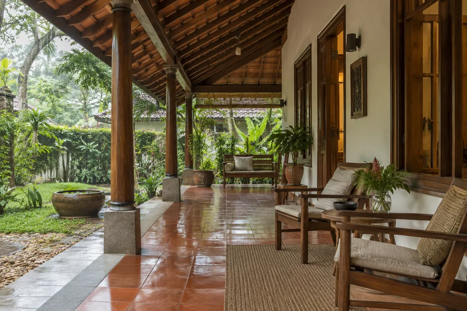 AI-generated concept: Kerala-inspired shaded veranda, teak outdoor chairs, terracotta floor, timber roof supports, lush garden, gentle monsoon daylight.